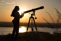 在夏季日落时透过望远镜看风景的年轻女子剪影