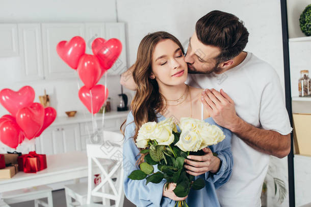 快乐的女孩拿着玫瑰花束闭着眼睛, 而男友亲吻脸颊和拥抱女朋友