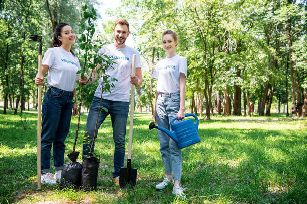 年轻的微笑的志愿者与新的树, 耙, 浇灌罐头并且铲站立在绿色公园