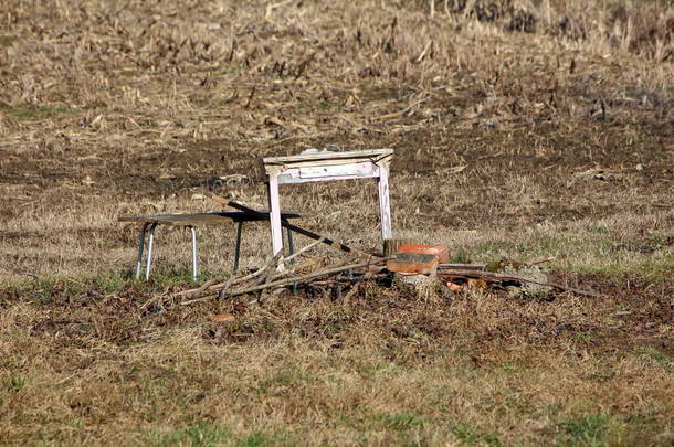 在温暖阳光明媚的冬日，在当地田园的玉米地旁边，有一个破旧的老式复古桌子，旁边有一个小抽屉，旁边有破碎的木制长椅，旁边有金属框架，周围有木桩和未切的干草