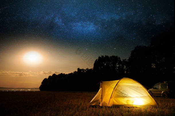 旅行和露营的概念 - 营地帐篷在夜晚的天空<strong>下</strong>满是星星。橙色照明帐篷和汽车。美丽的自然 - 田野，森林，平原。月亮和月光