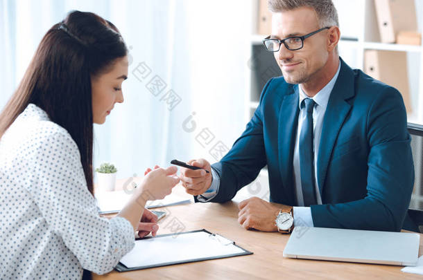 微笑的保险代理人给予笔给女性客户在桌面上签署文件在办公室