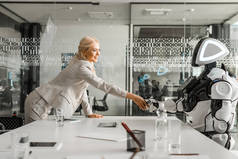 微笑的女商人和坐在会议室桌子前的机器人握手