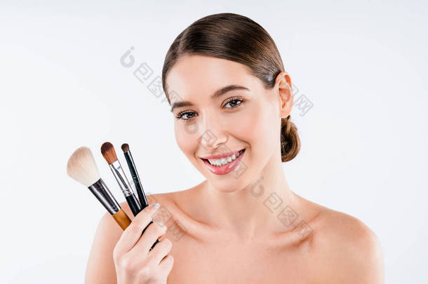 年轻女人有着洁白而明亮的皮肤，微笑着，拿着化妆品刷子，被白色的背景隔离开来。美容美发概念