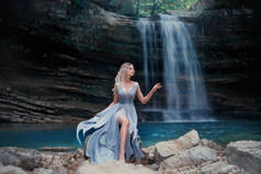 一个卷曲的金发女孩在一个豪华的蓝色礼服坐在白色的石头在一个神话般的景观的背景下。河美人鱼附近的湖与瀑布。艺术摄影