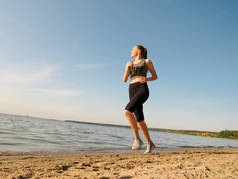在一个温暖的阳光明媚的秋日里，一位身材苗条的年轻女子穿着黑色训练紧身衣和一件白色T恤衫，沿着河边奔跑。户外运动、积极生活方式、运动和健康跑步的概念