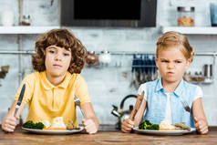 可爱的不快乐的孩子拿着餐具, 看着相机, 而坐在餐桌上吃蔬菜