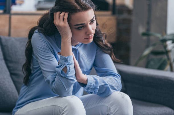悲伤的女人在蓝色的衬衫坐在灰色的沙发上, 看着在家里的距离, 悲伤的混乱的概念