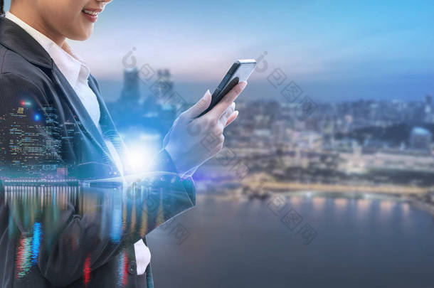 年轻的女商人使用手机与现代城市建筑背景。未来电信技术与物联网概念.