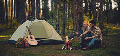 年轻的一对游客正在探索新的地方。有魅力的女人和英俊的男人都在大自然上呆着。坐在篝火和森林中的旅游帐篷附近, 制作棉花糖.