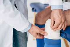 在足球运动员腿上系弹性绷带的理疗师在白色外套的局部视图