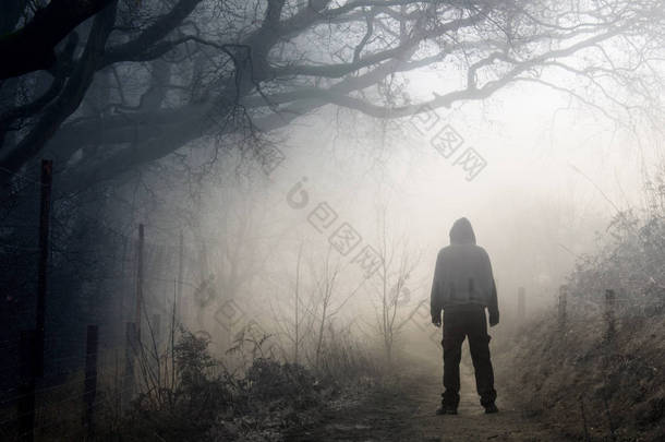 An artistic double exposure of a lone hooded figure on a path in the countryside on a spooky, foggy 