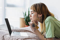 头戴信用卡的纹身妇女躺在床上看笔记本电脑的侧视图