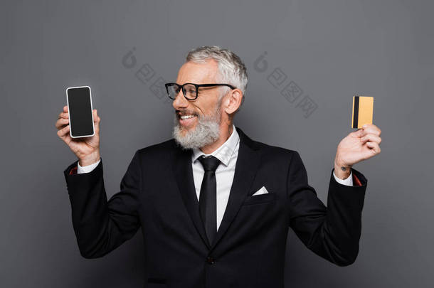 快乐的中年商人持有信用卡和智能手机，灰色屏幕上的空白屏幕