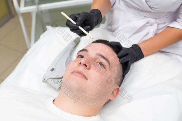 一个年轻英俊的男人躺在皮肤科办公室的沙发上的画像，而一个戴着黑色医疗手套的美容师的手拿着一支白色的化妆品铅笔，在他的脸上做记号。