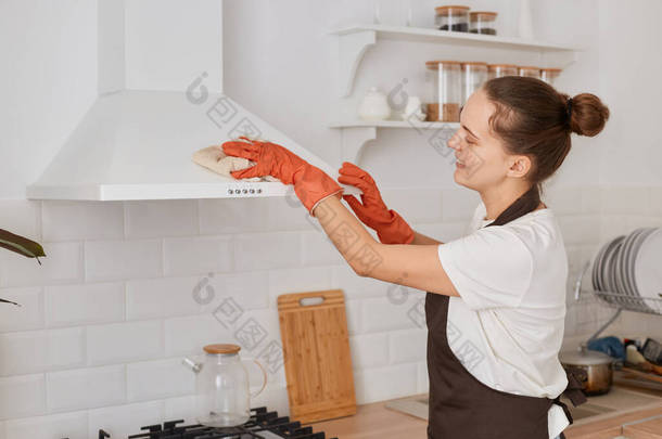 在家里厨房里，一个穿着白色T恤、棕色围裙和橙色手套，头戴发髻的女管家在用抹布和电动清洁罩洗碗时的水平照.