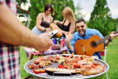 人类的手拿着一个烧烤钳和一个多汁美味的肉牛排，以及一个烧烤烤肉和蔬菜的背景和一群野餐的朋友在一起，他们都很开心。