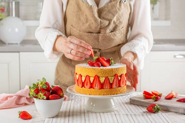 穿围裙做饭的女人在家里厨房里做法式蛋糕。用热那亚海绵、外交奶油和新鲜草莓制成的水果蛋糕.
