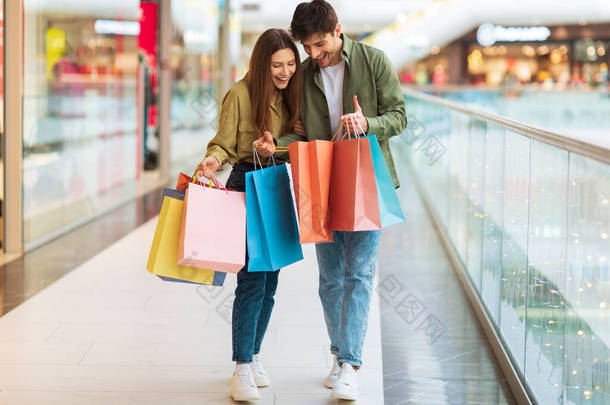 Excited Couple Looking Into Shopping Bags Standing In Modern Mall