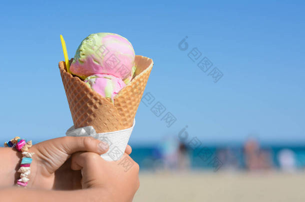 夏日阳光明媚的海滩上，小女孩拿着华夫饼筒和一勺美味的彩色冰淇淋，特写。案文的篇幅