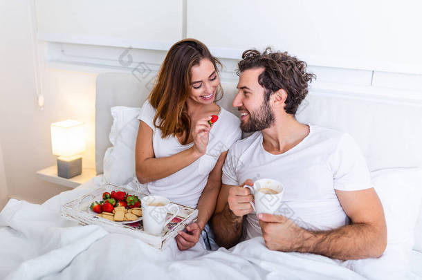 可爱的夫妇在卧室的床上吃早餐。漂亮的女人在床上喂男朋友草莓，在卧室里吃早餐和咖啡