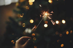 新年快乐！在黑暗的房间里，用女性的手在圣诞树下燃起火花。大气庆祝活动。手拿着烟火，照亮着装饰华丽的树木