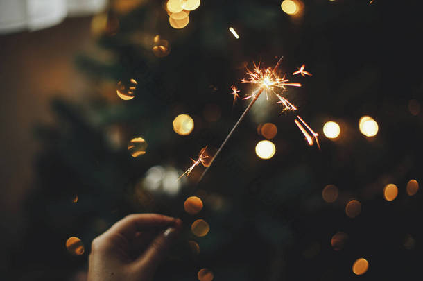 新年快乐！在黑暗的房间里，用女性的手在圣诞树下燃<strong>起火</strong>花。大气庆祝活动。手拿着烟火，照亮着装饰华丽的树木