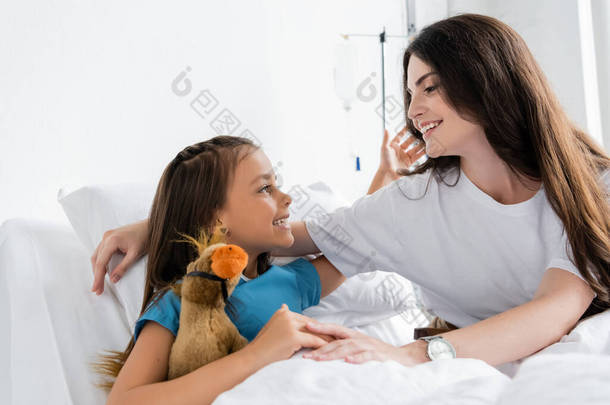 积极的孩子抱着妈妈，在诊所的床上抱着柔软的玩具