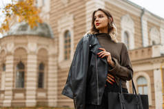 秋天的城市里，穿着时髦的黑色皮夹克，穿着绿色毛衣，提着包走在一座古色古香的建筑前，美丽而时尚的年轻姑娘