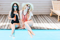 美丽的年轻妇女在泳装喝夏天饮料和微笑在照相机附近水池