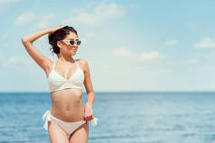 迷人的年轻女子在度假村附近的海边戴墨镜和白色泳装