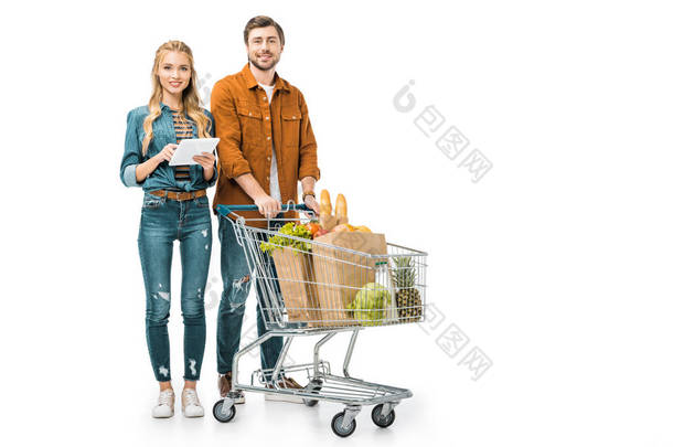 快乐的女孩检查购物清单上的数字平板电脑, 而她的男朋友站在附近与推车满纸袋与产品查出白色