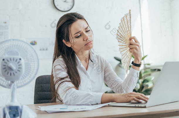 疲惫的女商人使用笔记本电脑, 而空调空气与电风扇和手风扇在办公室