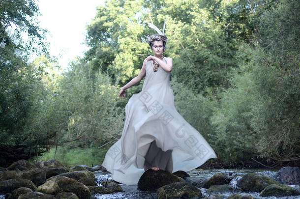 穿着灰色<strong>真丝</strong>连衣裙、羽毛头饰和海蜗牛贝壳项链的年轻女子站在一条暴风雨的河水流中的岩石上, 波浪着她的衣服的火车。