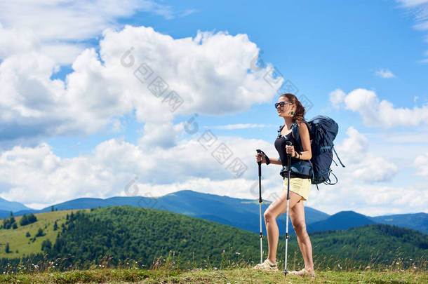 年轻女子背包徒步登山小径, 行走在草地上的山丘上, 穿着背包和太阳镜, 使用徒步棍, 享受夏日阳光明媚的一天在山上。旅游理念