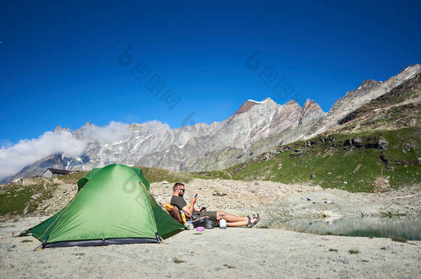 从侧面看男性游客坐在有雄伟岩石山背景的旅游帐篷旁.人们使用智能手机，在蓝天下的群山中享受着休息。旅行、远足和露营的概念