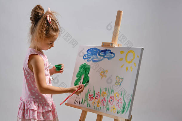 身穿粉色连衣裙的小女孩艺术家站在画架后面，画上画笔，与白色画室背景隔离。中特写镜头.