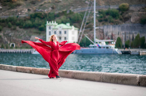 一个穿着红色连衣裙的女人，一个穿着<strong>飘逸</strong>的长丝翅膀的时装模特，在堤岸上飘扬着面料.