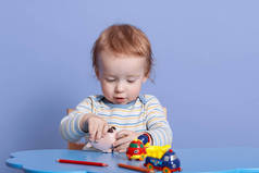 小白种男孩的近照。可爱的婴儿坐在孩子的办公桌前，一个人玩玩具。可爱的孩子快乐而兴奋，穿着脱光的衬衫，被蓝色的背带隔开.
