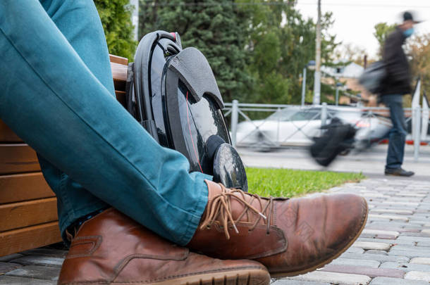 电动汽车，创新的私人汽车，自我平衡的电动汽车，未来的生态城市交通，靠近一个穿着牛仔裤和棕色鞋子的男人坐在城市公园的长椅上