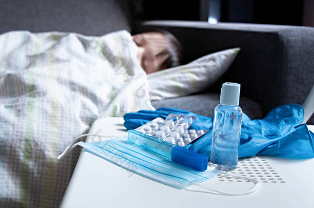 生病的孩子躺在床上，毯子盖住了他的身体。在平板电脑、保护膜、温度计、手套和瓶子手部清洁剂旁边的椅子上