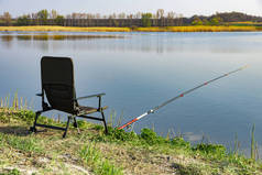 早上湖滨上有一张空椅子和一根钓竿。 绿草在大湖畔，蓝天，清早时分