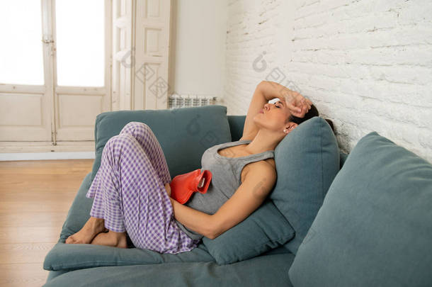 年轻美丽的拉丁裔妇女在<strong>痛苦</strong>的表达遭受胃痉挛和月经期间<strong>痛苦</strong>拿着一个热水瓶对腹部躺在沙发上。妇女健康概念