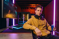 Portrait of young serious handsome pro gamer sitting at desk with gaming equipment, holding energy d