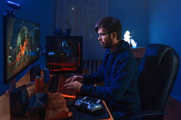 专注的职业游戏玩家游戏，参加在线竞赛，在家坐在电脑前。低光，霓虹灯色，侧视。专业的网络游戏玩家在电脑上进行在线游戏训练