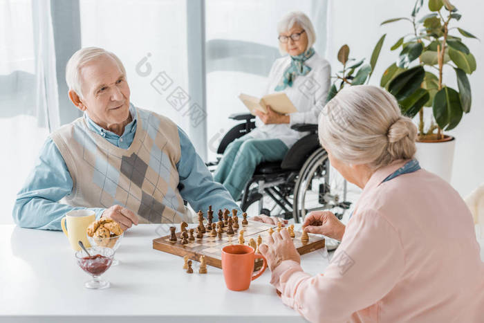 高级夫妇下棋, 而高级妇女坐在轮椅上看书养老