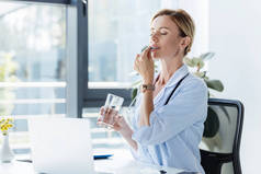成年女医生在白色大衣拿着玻璃杯水, 并在桌子上用笔记本电脑在办公室里吃药丸