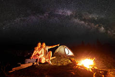 晚上在山上露营。旅游家庭休息在照明帐篷前和燃烧篝火。女子抱着小女孩, 男子指着明亮的星星在黑暗的天空和银河