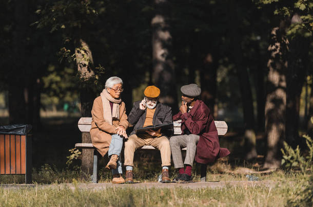 资深跨种族人士坐在好友身边，带着相册，在公园里用智能手机交谈