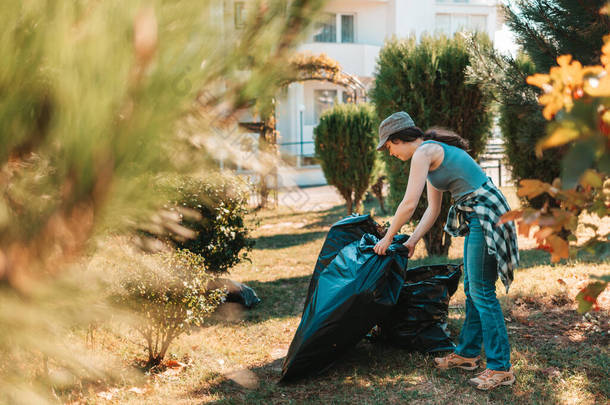 环境污染和地球日的概念。妇女自愿在公园的房子前面做清扫工作。她把垃圾袋堆在一起了。<strong>靠近点</strong>.